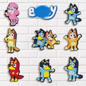 May include: A collection of colorful cartoon character magnets, including a pink poodle, various dog characters, and a blue word. The magnets are arranged on a white brick wall.