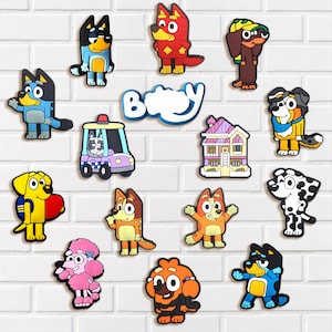 May include: A collection of colorful cartoon character magnets, including dogs, a house, and a police vehicle. The magnets are in various colors, such as blue, red, yellow, and orange, and are arranged on a white brick wall.