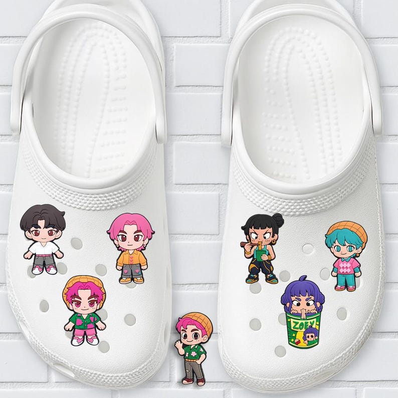 K&ouml;nnte beinhalten: Wei&szlig;e Clogs, verziert mit Cartoon-Charms. Die Charms zeigen verschiedene Charaktere mit unterschiedlichen Haarfarben und Outfits. Die Clogs befinden sich auf wei&szlig;em Hintergrund.
