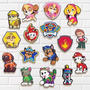May include: A collection of colorful Paw Patrol character charms in various shapes. The charms feature cartoon dogs in uniforms and badges, including Skye, Chase, Marshall, and Rubble. The charms are made of a glossy material and are arranged on a white brick wall.
