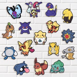 May include: A collection of colorful cartoon character charms, including a butterfly, a purple snake, and a yellow and black bear-like figure. The charms are arranged on a white brick wall background.
