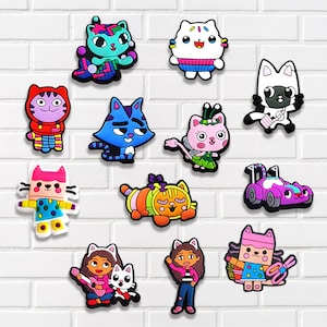 May include: A collection of colorful cat-themed charms, featuring various designs and characters. The charms include cats in costumes, a car, and characters with musical instruments. The charms are made of a glossy material and are set against a white brick wall.