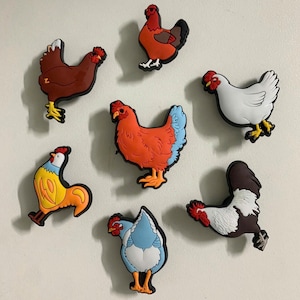May include: A collection of seven colorful chicken-shaped decorative magnets. The magnets feature various colors, including red, brown, white, blue, and yellow, with black outlines. The chickens are in different poses.