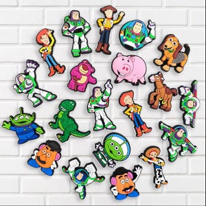May include: A collection of colorful Toy Story character charms, including Woody, Buzz Lightyear, and others, arranged on a white brick background. The charms feature vibrant colors and detailed designs, perfect for fans of the animated film.