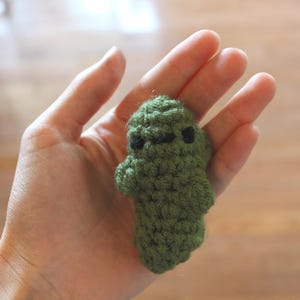 May include: A small, handmade green crochet creature with black eyes. The plush toy is held in an open hand, showcasing its textured, detailed design. The item is a unique, handcrafted item.