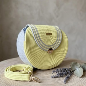 May include: A round, yellow and gray rope handbag with a matching shoulder strap. The bag has a gold-colored clasp and a small rectangular tag. The bag is sitting on a wooden surface.