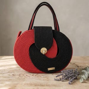 May include: A round handbag with a unique design, featuring a combination of red and black rope. The bag has a flap closure with a gold-colored clasp and two black handles with red accents. A small gold tag is visible.