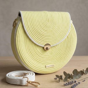 May include: A yellow, circular handbag made of coiled rope, with a white trim and a gold clasp. The bag has a matching white strap with gold hardware. The label reads "Yaya & Pimpi".
