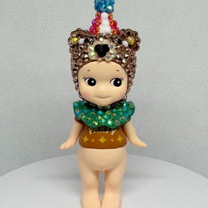 May include: A small, nude-colored figurine with a detailed, jeweled headdress and collar. The headdress is brown with a black heart-shaped accent and a colorful, beaded top. The figurine wears a brown shirt with a green collar and gold accents.