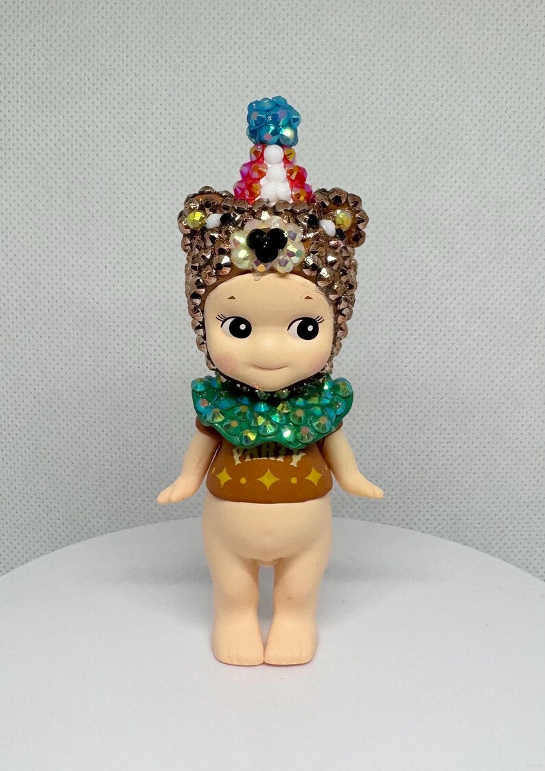 May include: A small, nude-colored figurine with a detailed, jeweled headdress and collar. The headdress is brown with a black heart-shaped accent and a colorful, beaded top. The figurine wears a brown shirt with a green collar and gold accents.