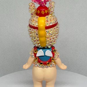 May include: A decorative figurine with a light skin tone, adorned with iridescent rhinestones. It features a pink, flower-like headpiece, a red and yellow crown, and a blue and red collar with white wing accents. The figurine has a yellow, curved back piece.