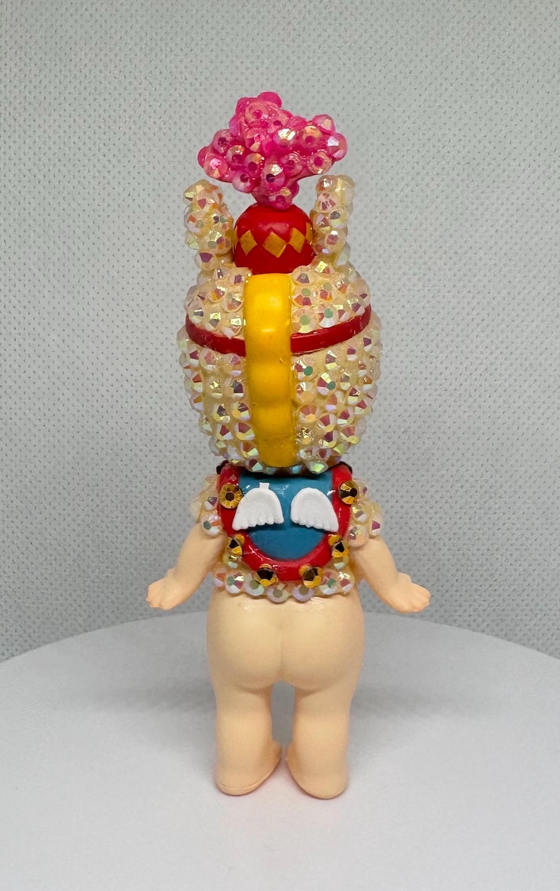 May include: A decorative figurine with a light skin tone, adorned with iridescent rhinestones. It features a pink, flower-like headpiece, a red and yellow crown, and a blue and red collar with white wing accents. The figurine has a yellow, curved back piece.