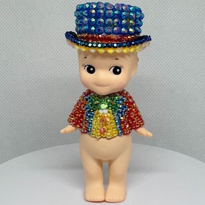 May include: A small doll with a round head and a painted face, wearing a blue jeweled top hat and a rainbow-colored jacket. The doll has a light skin tone and is standing on a white surface.