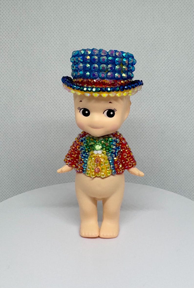 May include: A small doll with a round head and a painted face, wearing a blue jeweled top hat and a rainbow-colored jacket. The doll has a light skin tone and is standing on a white surface.