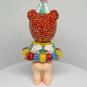 May include: A whimsical figurine of a bear-like character, adorned with sparkling red and gold embellishments. It wears a colorful tutu and a party hat with a pink pom-pom. The figurine is standing on a white surface.