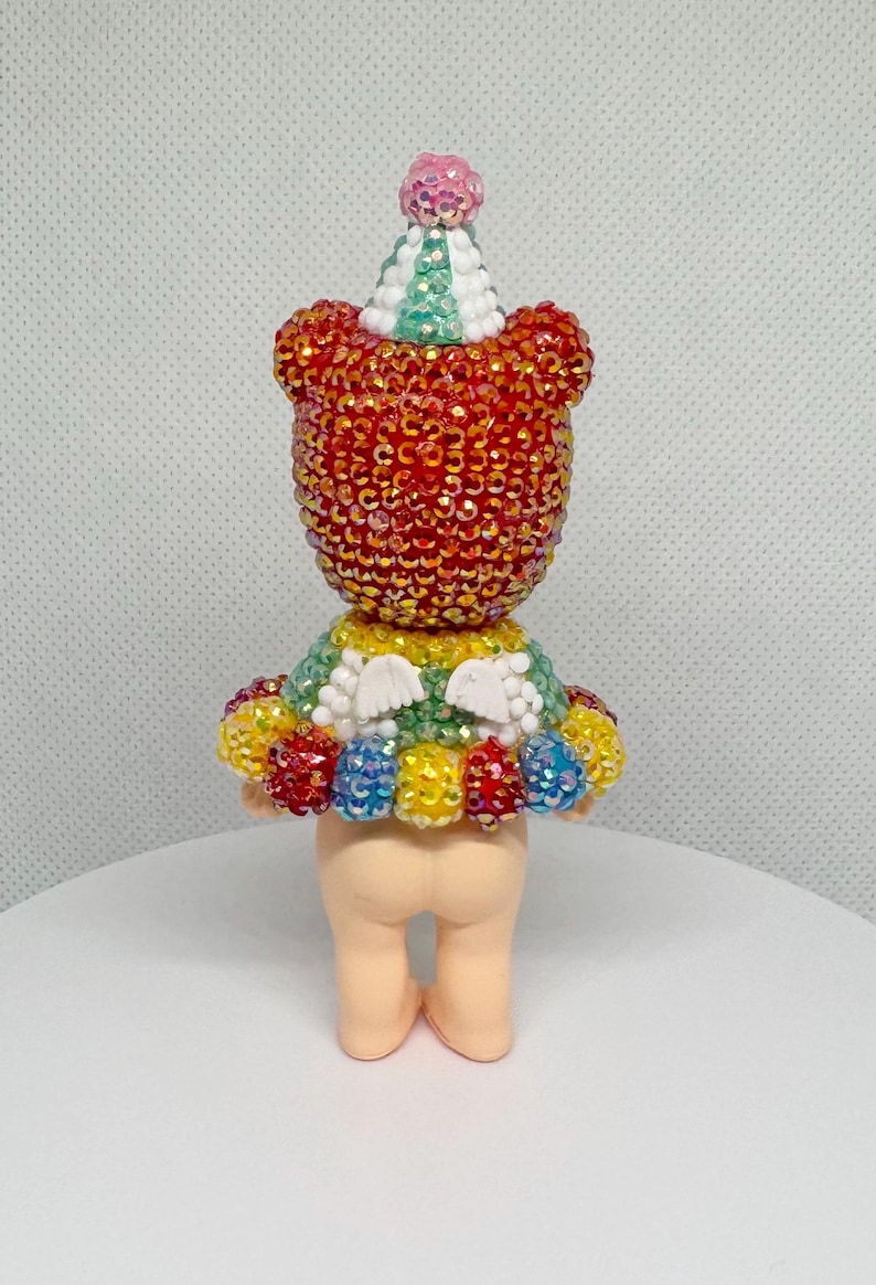 May include: A whimsical figurine of a bear-like character, adorned with sparkling red and gold embellishments. It wears a colorful tutu and a party hat with a pink pom-pom. The figurine is standing on a white surface.