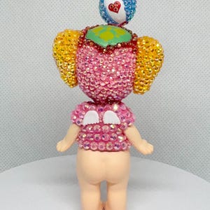 May include: A whimsical figurine with a pink, gem-studded torso and head, adorned with yellow and blue accents. The figure has small arms and legs, and a small, heart-shaped detail on the head. The overall design is playful and colorful.