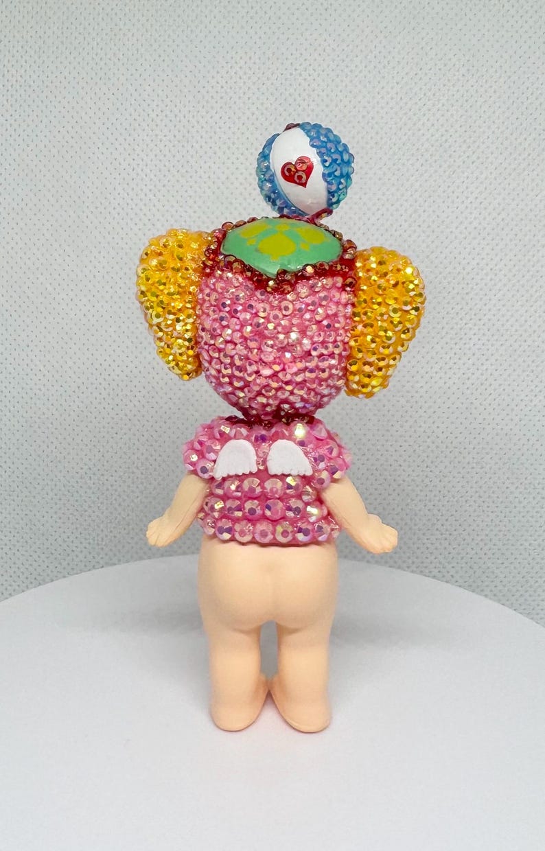 May include: A whimsical figurine with a pink, gem-studded torso and head, adorned with yellow and blue accents. The figure has small arms and legs, and a small, heart-shaped detail on the head. The overall design is playful and colorful.