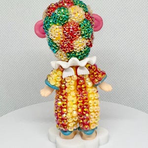 May include: A decorative figurine of a clown with a colorful, jeweled head and costume. The clown has pink ears, a white ruffled collar, and small arms. The costume is yellow, red, and orange with blue accents. The figurine is standing on a white cloud-shaped base.