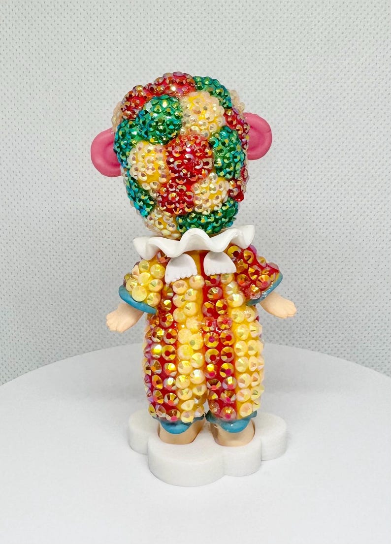 May include: A decorative figurine of a clown with a colorful, jeweled head and costume. The clown has pink ears, a white ruffled collar, and small arms. The costume is yellow, red, and orange with blue accents. The figurine is standing on a white cloud-shaped base.