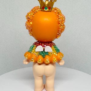May include: A whimsical figurine with an orange head and a gold crown. The figure has a beaded orange halo and a colorful beaded collar. The base of the figure is a light beige color.