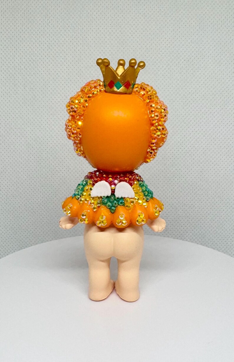 May include: A whimsical figurine with an orange head and a gold crown. The figure has a beaded orange halo and a colorful beaded collar. The base of the figure is a light beige color.