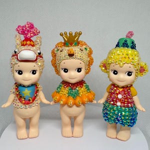 May include: Three small, decorative figurines with detailed costumes. The figurines have large eyes and are adorned with sparkling embellishments. One wears a lion headdress, another a circus-themed hat, and the third a crown.