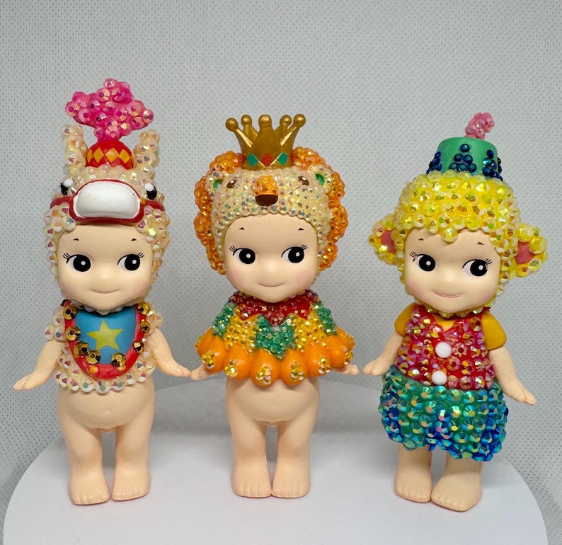 May include: Three small, decorative figurines with detailed costumes. The figurines have large eyes and are adorned with sparkling embellishments. One wears a lion headdress, another a circus-themed hat, and the third a crown.