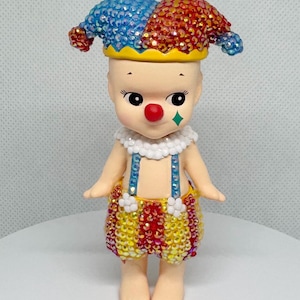 May include: A whimsical figurine of a clown with a red nose, a green diamond-shaped cheek mark, and a jester's hat. The hat is split into blue and red sections, and the clown's outfit is adorned with colorful embellishments. The figurine has a cheerful expression.