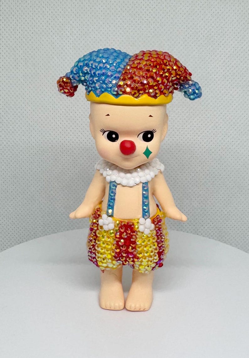May include: A whimsical figurine of a clown with a red nose, a green diamond-shaped cheek mark, and a jester's hat. The hat is split into blue and red sections, and the clown's outfit is adorned with colorful embellishments. The figurine has a cheerful expression.