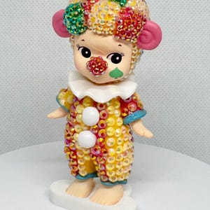 May include: A whimsical figurine of a clown with a colorful, sequined outfit. The clown has a white ruffled collar, a red nose, and a green cheek. The clown's hair is a mix of colorful sequins. The figurine stands on a white base.
