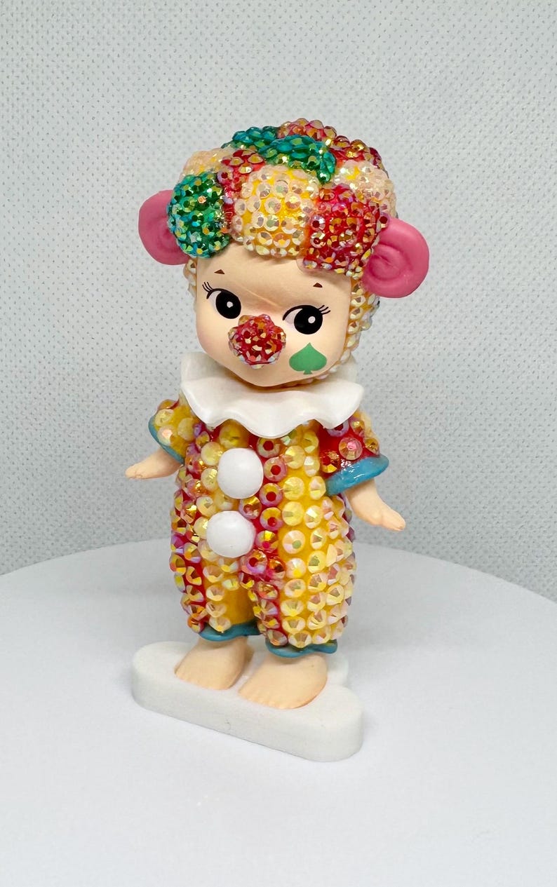 May include: A whimsical figurine of a clown with a colorful, sequined outfit. The clown has a white ruffled collar, a red nose, and a green cheek. The clown's hair is a mix of colorful sequins. The figurine stands on a white base.