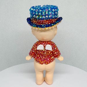 May include: A small figurine wearing a blue jeweled top hat and a red jeweled shirt with white accents. The figurine has a light skin tone and is standing upright.