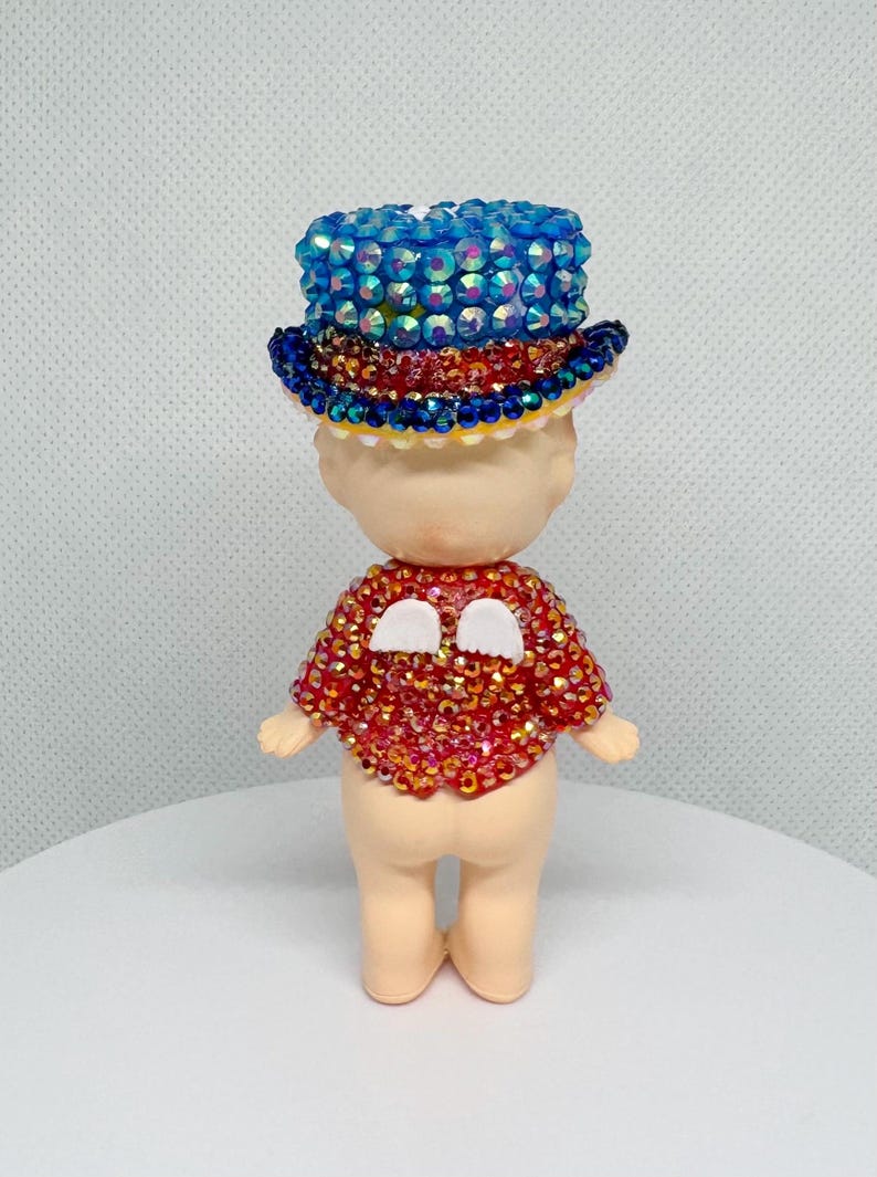 May include: A small figurine wearing a blue jeweled top hat and a red jeweled shirt with white accents. The figurine has a light skin tone and is standing upright.