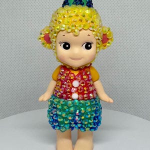 May include: A whimsical figurine of a doll-like character with a yellow sequined head and ears, wearing a colorful sequined vest and skirt. The figurine has a green hat with blue accents and a pink flower on top. The doll is standing on a white surface.