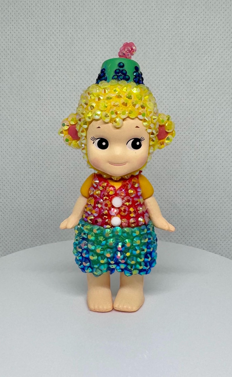 May include: A whimsical figurine of a doll-like character with a yellow sequined head and ears, wearing a colorful sequined vest and skirt. The figurine has a green hat with blue accents and a pink flower on top. The doll is standing on a white surface.