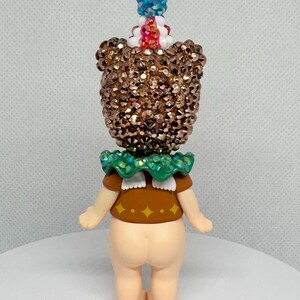 May include: A whimsical figurine with a sequined head and a green ruffled collar. The figure has a brown top with yellow diamond accents and a light skin tone. A blue and red gem adorns the top of the head.
