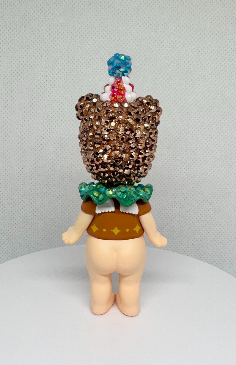 May include: A whimsical figurine with a sequined head and a green ruffled collar. The figure has a brown top with yellow diamond accents and a light skin tone. A blue and red gem adorns the top of the head.