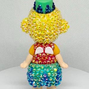 May include: A whimsical figurine with a yellow, gem-studded head and body, a green hat, and a colorful skirt. The figurine has small white wings and is posed with arms at its sides. The figure is standing on a white surface.