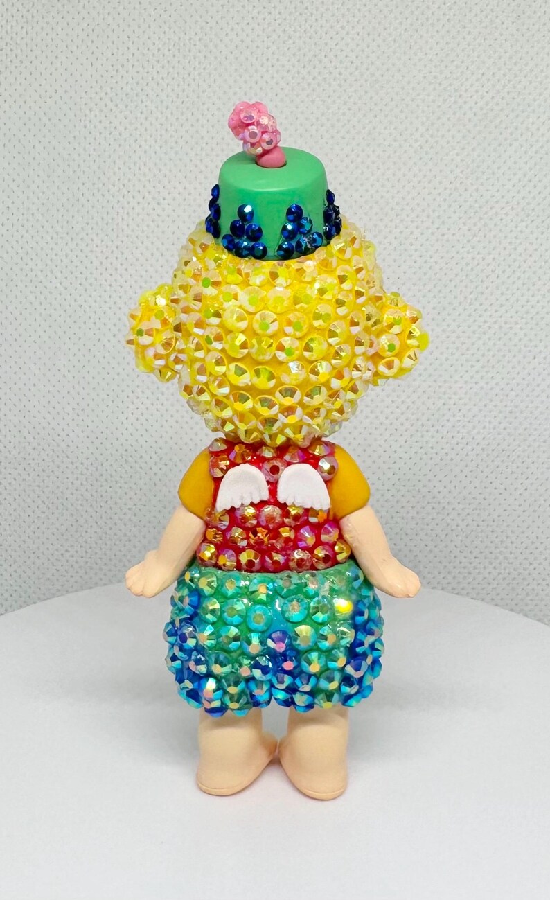 May include: A whimsical figurine with a yellow, gem-studded head and body, a green hat, and a colorful skirt. The figurine has small white wings and is posed with arms at its sides. The figure is standing on a white surface.