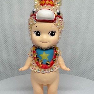 May include: A small, doll-like figurine with a whimsical design. It features a light skin tone, large eyes, and a smiling expression. The figurine is adorned with a decorative headpiece, a bib with a star, and a collar with gold accents. The overall aesthetic is playful and collectible.