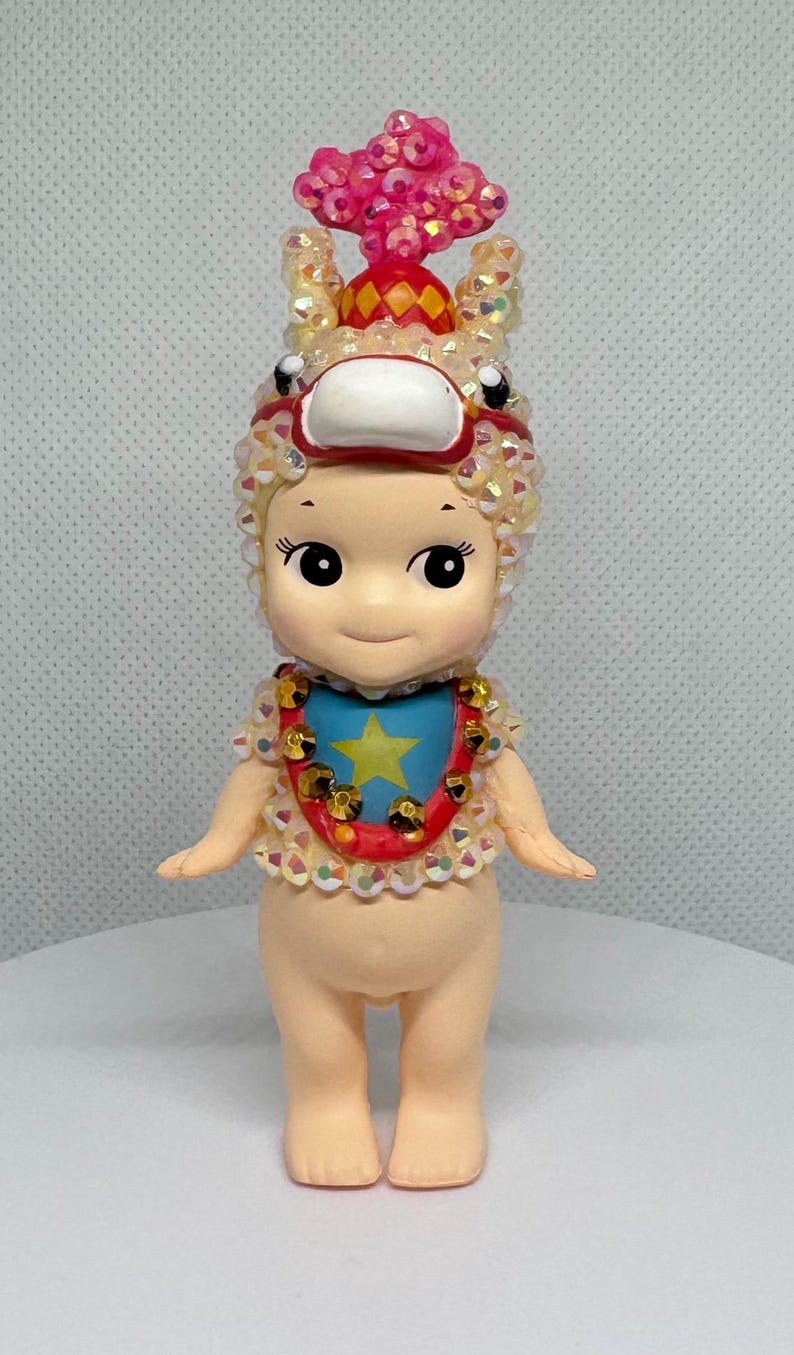 May include: A small, doll-like figurine with a whimsical design. It features a light skin tone, large eyes, and a smiling expression. The figurine is adorned with a decorative headpiece, a bib with a star, and a collar with gold accents. The overall aesthetic is playful and collectible.