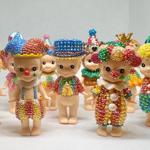 May include: A collection of small, decorative figurines. Each figurine is adorned with colorful, sparkling embellishments, and dressed in unique outfits, including clown and animal-themed costumes. The figurines have a whimsical and playful appearance.