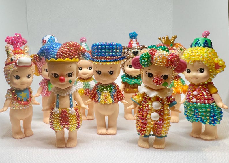 May include: A collection of small, decorative figurines. Each figurine is adorned with colorful, sparkling embellishments, and dressed in unique outfits, including clown and animal-themed costumes. The figurines have a whimsical and playful appearance.