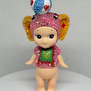 May include: A whimsical figurine of a cherubic character adorned with a pink and gold jeweled elephant-ear hat and matching top. A decorative ball sits atop the head. The figurine has a green shirt and is standing on a white surface.
