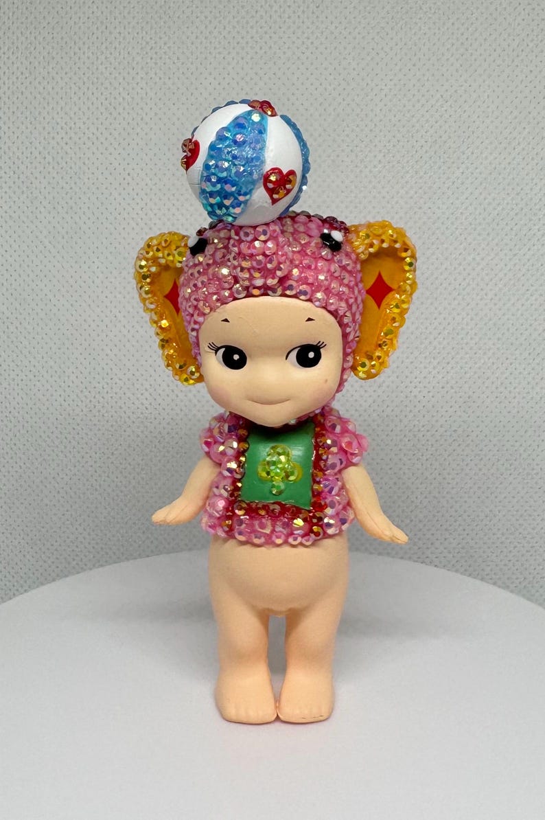May include: A whimsical figurine of a cherubic character adorned with a pink and gold jeweled elephant-ear hat and matching top. A decorative ball sits atop the head. The figurine has a green shirt and is standing on a white surface.