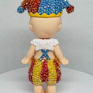 May include: A whimsical figurine of a jester, adorned with a red and blue jeweled hat, a white collar, and yellow, red, and gold shorts. The figure has small white wings and a blue cross design on the back.