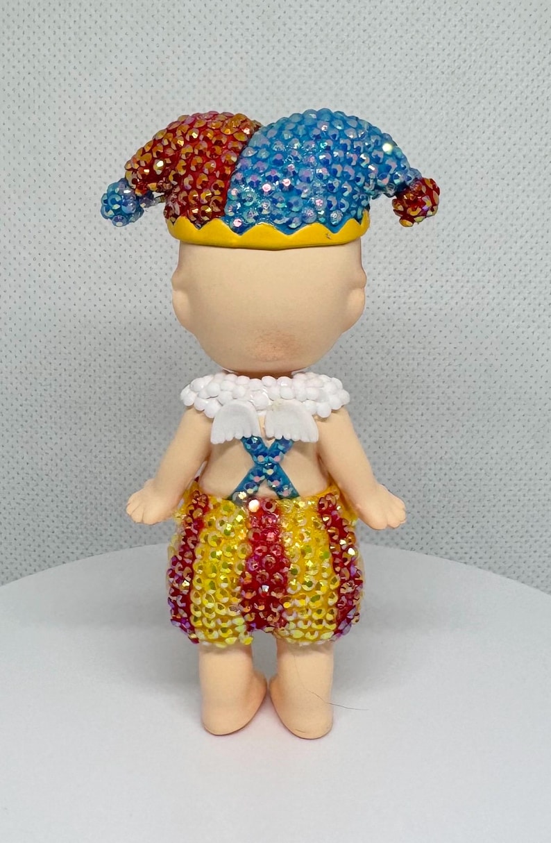 May include: A whimsical figurine of a jester, adorned with a red and blue jeweled hat, a white collar, and yellow, red, and gold shorts. The figure has small white wings and a blue cross design on the back.