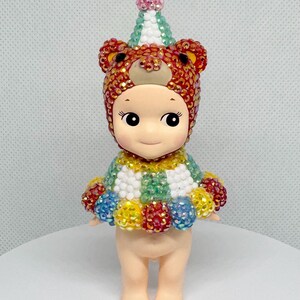 May include: A small figurine of a doll wearing a bear-themed hat and a colorful outfit. The hat is red and gold with bear ears. The doll's outfit is a mix of colors, including green, yellow, blue, and red. The doll has a cute face.