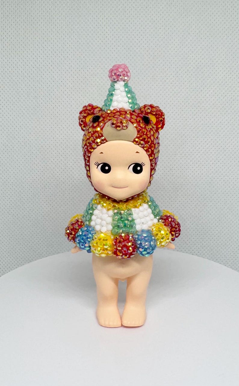 May include: A small figurine of a doll wearing a bear-themed hat and a colorful outfit. The hat is red and gold with bear ears. The doll's outfit is a mix of colors, including green, yellow, blue, and red. The doll has a cute face.